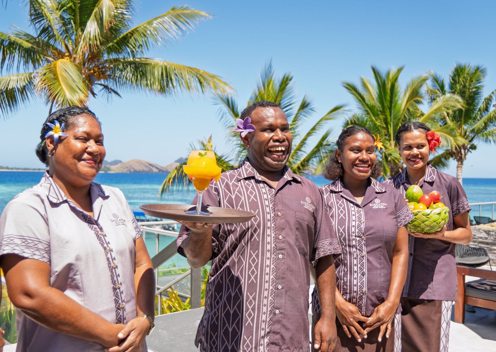 Smiling resort staff in island uniforms holding a cocktail and a basket of fruit by the sea