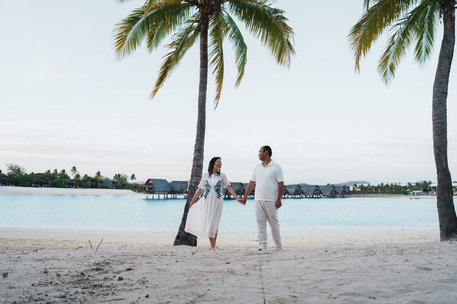 Couple holding hands beneath a palm tree with overwater bures behind