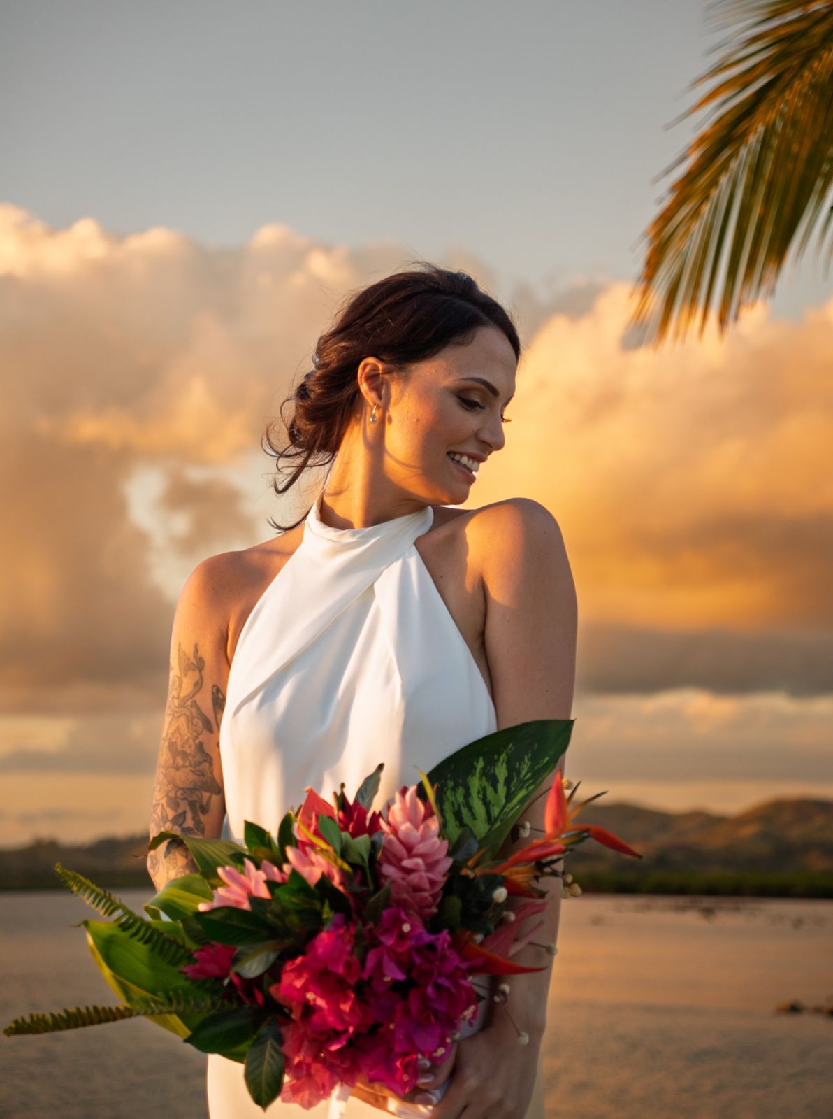 Bride smiling over her shoulder holding a tropical bouquet against a golden sky
