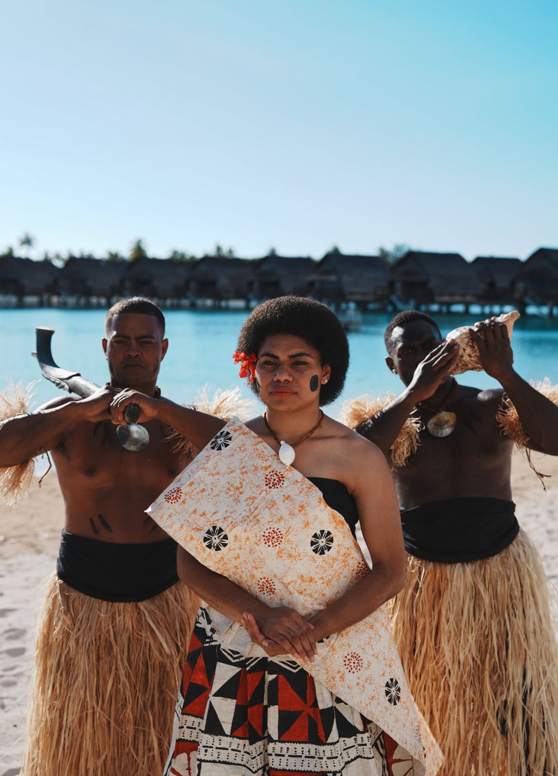 Three Fijians in traditional dress with a club and conch shell by the lagoon