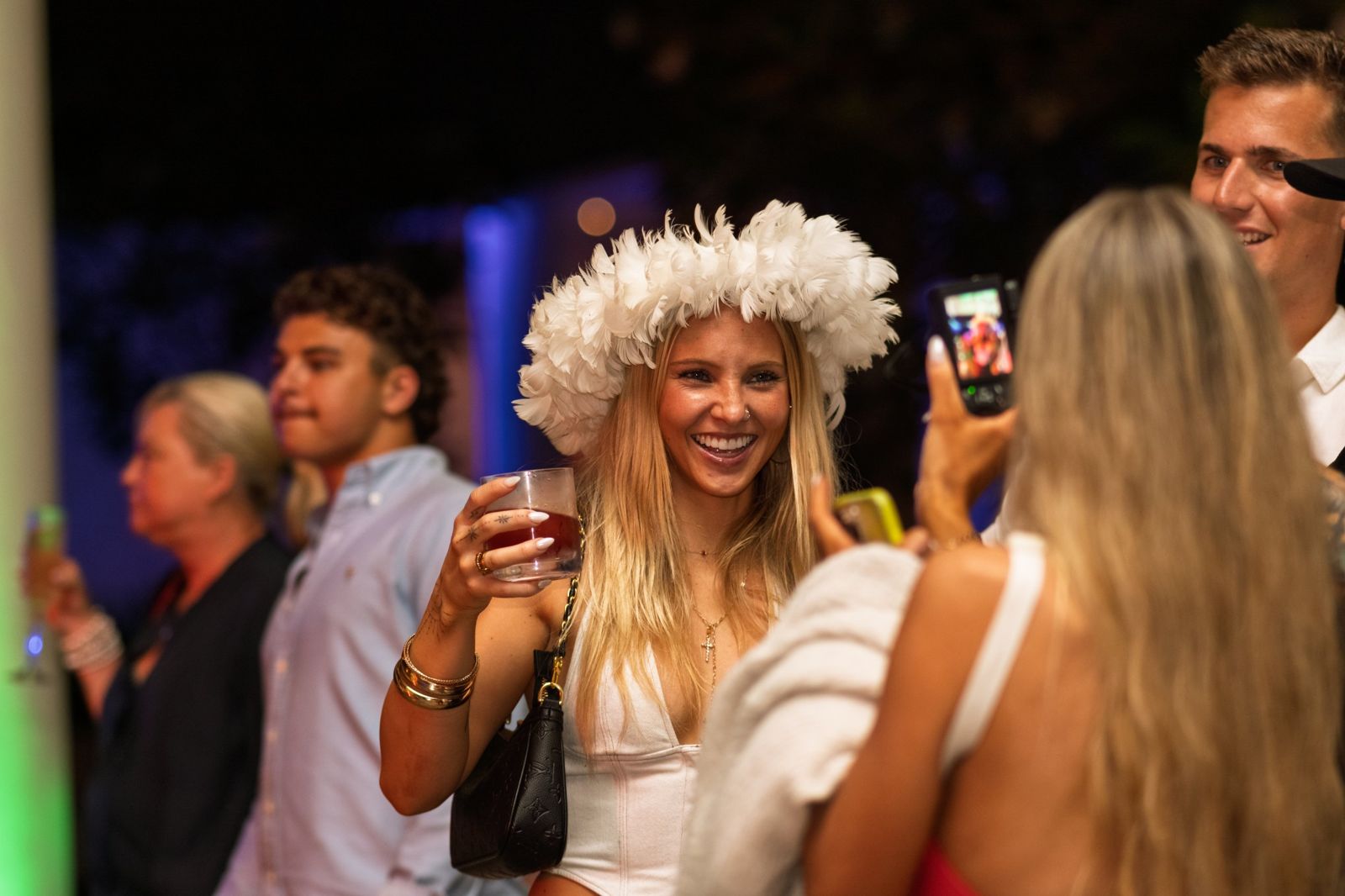 Guest in a white feather crown laughing with a drink at a night party