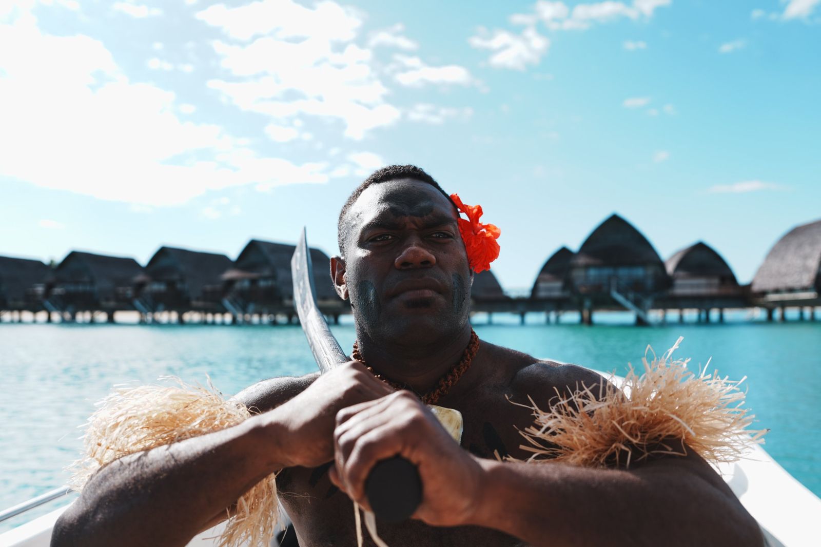 Fijian warrior with a carved club in front of overwater bures